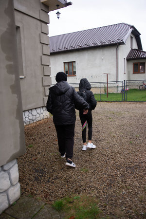 Two figures are seen from the back, walking on a gravel path. They're dressed in warm jackets, suggesting a chilly day. A house is visible in the background, setting the scene in a residential area.の写真素材