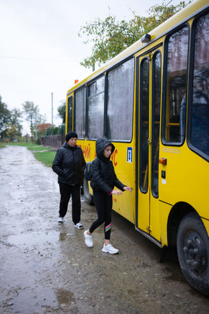 This image shows a grandmother walking alongside a young girl in a hooded jacket as she gets on a yellow bus on a cloudy day. The pair are on a wet, rural roadside.の写真素材