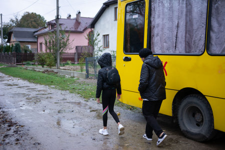 On a cloudy day, two figures, one appearing younger, are walking alongside a bright yellow bus. The road is muddy, and houses form a backdrop to the scene, creating a realistic context.の写真素材