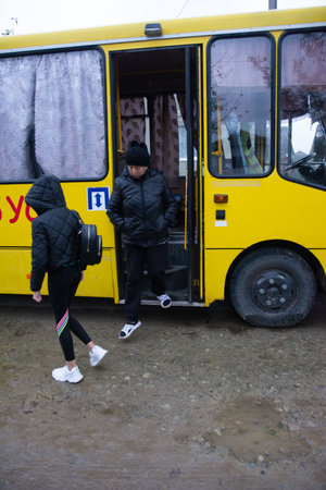 An older woman exits a yellow bus, wearing a dark coat and hat. A younger girl in leggings and a puffy jacket walks in front of the bus. Ground is wet suggesting a rural setting.の写真素材
