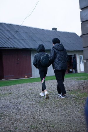 In this outdoor shot, two figures in quilted jackets walk side-by-side on a gravel path, with a low building in the background, under an overcast sky, evoking a sense of companionship.の写真素材