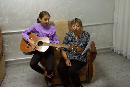 A young girl practices playing the guitar while her instructor looks at a sheet of music. They are sitting in a cozy room with neutral decor, creating a focused learning environment.の写真素材