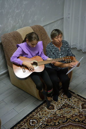 A young girl plays an acoustic guitar while sitting next to an older woman, presumably her grandmother, who holds sheet music. They appear to be learning or practicing music together indoors.の写真素材