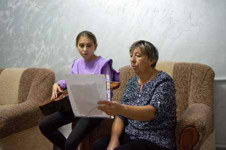 A young girl sits with an older woman, possibly her grandmother, both holding sheet music. The girl plays a stringed instrument in a casual, indoor setting, focused on the music.の写真素材