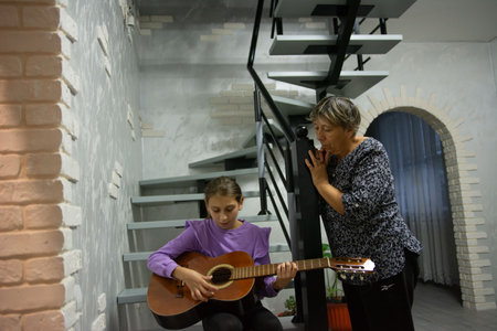 In a modern home, a young girl is seated and passionately playing a guitar, while an older woman stands nearby attentively watching and listening.の写真素材