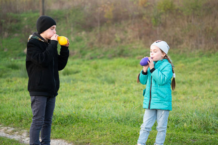 Children create joyful sounds by blowing colorful balloons in a vibrant green field, showcasing their playful spirit under the warm sun and a clear blue sky.の写真素材