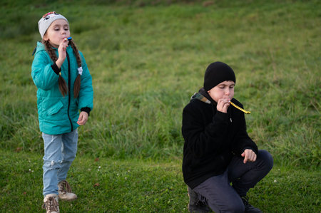 Two children engage in playful excitement, surrounded by vibrant greenery while blowing brightly colored noisemakers on a sunny afternoon, expressing pure joy and creativity.の写真素材