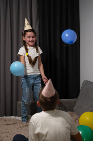 Two cheerful children are engaged in a playful atmosphere filled with balloons and party hats, celebrating a birthday together in a cozy living room filled with joy.の写真素材