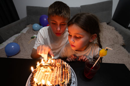 Two children lean in close, eyes bright with excitement as they prepare to blow out the candles on a delicious birthday cake, surrounded by festive decorations.の写真素材