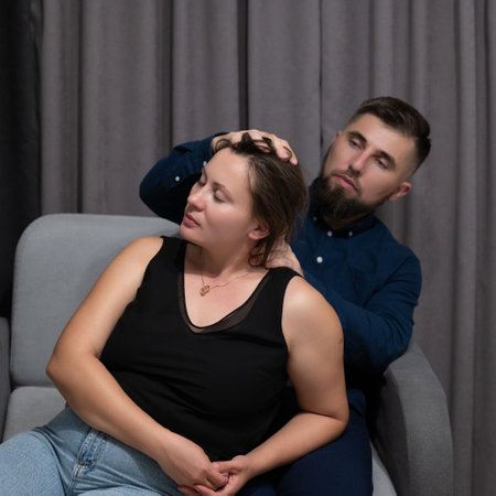 In a cozy living room, a man gently massages a woman's neck while she savors the relaxation, surrounded by soft lighting and warm decor that enhance the tranquil vibe.の写真素材