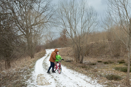 A child in a bright green jacket learns to ride a pink bicycle on a snow-covered trail, while an encouraging parent assists in a serene winter landscape.の写真素材