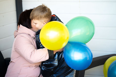 A moment of love as a child hugs a parent while holding vibrant balloons, capturing the essence of joy and warmth in a cozy outdoor space under the sun.の写真素材