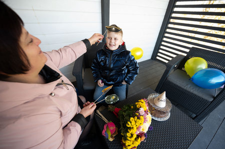 Delight fills the air as a woman lovingly places a pilot hat on a smiling child's head, celebrating a joyful birthday on a cozy porch adorned with colorful balloons.の写真素材