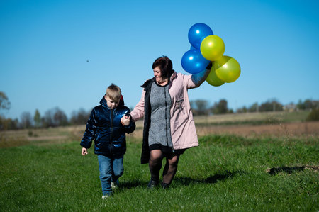 A mother and her son stroll hand in hand through lush green fields, celebrating a clear blue sky and vibrant balloons, embodying pure joy and connection.の写真素材