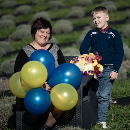 A woman seated comfortably holds a cluster of vibrant blue and yellow balloons while a young boy stands nearby, grinning and showcasing a beautiful bouquet of flowers.の写真素材