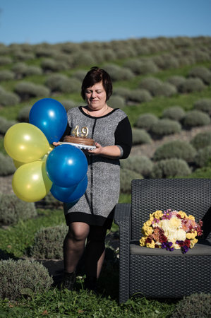 A woman holds a colorful bouquet of balloons and a birthday cake while walking through a scenic lavender field, celebrating her special day in a serene outdoor setting.の写真素材