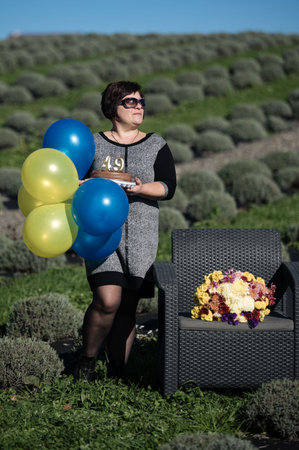 A woman stands in a picturesque lavender field, holding yellow and blue balloons, celebrating her special day with a beautiful floral arrangement nearby.の写真素材