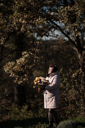 In a tranquil park, a woman admires a colorful bouquet of flowers, enveloped by the warm hues of autumn foliage, while soft sunlight bathes the scene in golden light.の写真素材