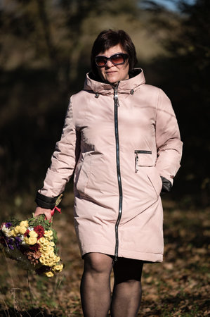 A woman strolls in a serene outdoor setting, dressed warmly in a fashionable coat while holding a colorful bouquet, embodying the joy of nature in autumn's embrace.の写真素材