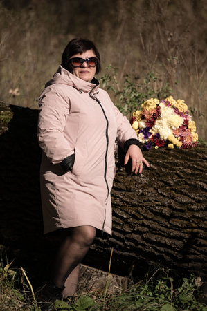 A confident woman enjoys the tranquility of nature, leaning on a large fallen tree, surrounded by colorful flowers that brighten the peaceful landscape.の写真素材
