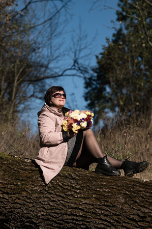 Sitting on a fallen tree, a woman in a light coat holds a vibrant bouquet, basking in the sunshine and the serenity of the surrounding landscape.の写真素材