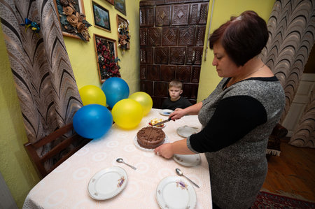 A warm celebration unfolds as a woman prepares to serve a chocolate cake, surrounded by cheerful balloons and a curious child, capturing a moment of happiness.の写真素材