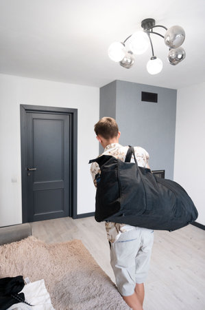 Male individual in casual attire stands in a modern living area, holding a large black bag on his shoulder, getting ready to head out into the day.の写真素材