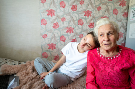 A young girl relaxes against her grandmother, both smiling softly while sitting on a plush bed covered with light fabric, enjoying quality time in a cheerful atmosphere.の写真素材
