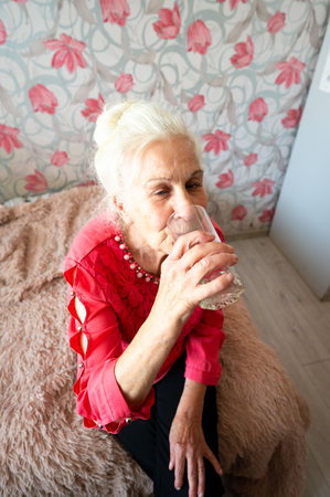An elderly woman savors a glass of water, reflecting tranquility and joy, while seated gracefully on a plush bed in a beautifully decorated space.の写真素材