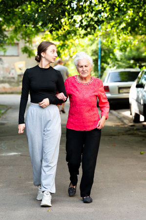 A young woman and an elderly lady stroll arm in arm, sharing smiles and laughter as they savor a sunlit afternoon in their peaceful residential area.の写真素材