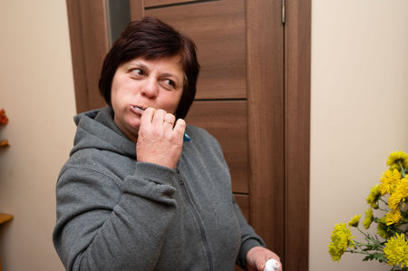 A woman in a gray hoodie stands by a wooden door, brushing her teeth while giving a thoughtful expression. Colorful flowers in a vase create a warm atmosphere.の写真素材