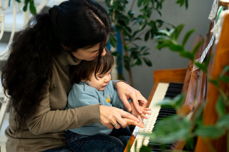A parent engages with their young child at a piano, guiding their tiny fingers over the keys, as sunlight bathes the cozy room adorned with plants and warmth of togethernessの写真素材