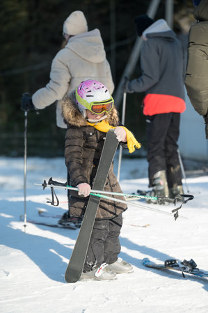 A young child stands on a snowy slope, equipped with skis and a colorful helmet, ready to join adults skiing nearby under the bright winter sun.の写真素材