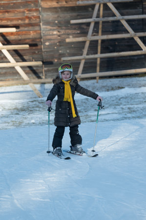 A cheerful young skier navigates the pristine snow, wearing a warm coat and bright scarf, surrounded by a scenic winter backdrop of wooden structures and sunshine.の写真素材