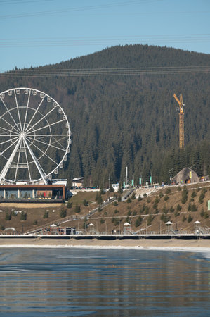 A giant Ferris wheel stands tall by a calm lake, surrounded by dense forested hills, capturing the joy of visitors enjoying a bright day in nature's beauty.の写真素材