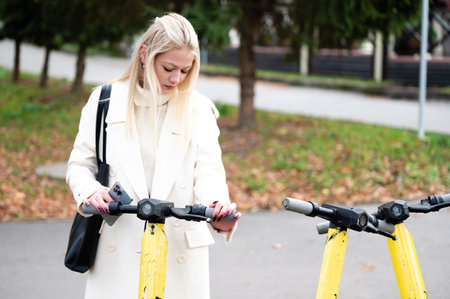 A young woman dressed in a chic coat inspects an electric scooter in a lively park, where the colorful fall foliage creates a picturesque backdrop for her outdoor adventure.の写真素材