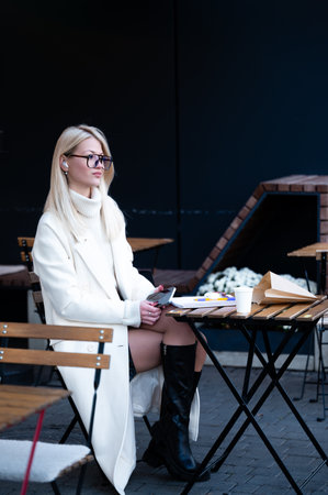 A fashionable woman sits at a rustic outdoor table, sipping coffee and absorbed in her work, surrounded by the charm of urban life on a crisp autumn day.の写真素材