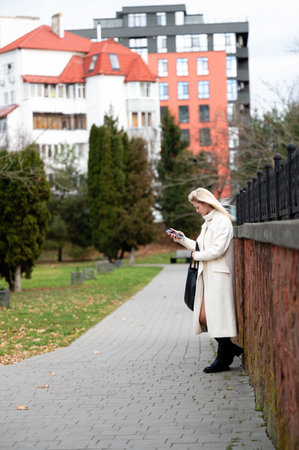 A stylish woman in a long coat stands on a walkway, focused on her phone, surrounded by vibrant autumn leaves and urban architecture on an overcast day.の写真素材