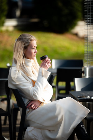 A young woman in a cozy white outfit savors her drink, unwinding at an outdoor cafe. Sunshine bathes the area, creating a peaceful atmosphere filled with nature.の写真素材