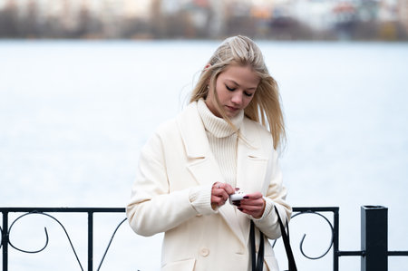 Standing by the lakeside, a woman in a white coat intently checks her phone. The soft breeze flows through her hair as she enjoys a peaceful afternoon in spring.の写真素材