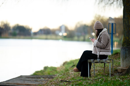Young woman nestled in cozy attire sits peacefully by the lake, immersed in her phone as golden hour light reflects on calm waters during a tranquil autumn evening.の写真素材