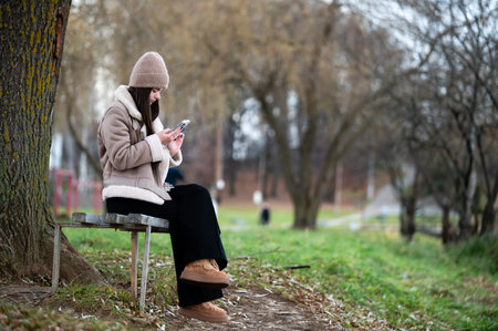 Sitting on a rustic bench, a young woman engages with her phone, surrounded by the tranquility of a winter park, highlighted by bare branches and subdued colors.の写真素材