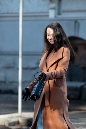 Walking through a city street, a fashionable woman with long hair proudly showcases her chic coat and handbag, radiating confidence and joy in the warm sunlight.の写真素材