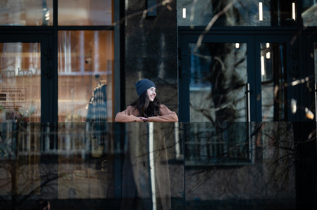A cheerful woman stands at a modern glass railing, smiling and looking out over a charming street scene filled with a warm ambiance as leaves drift gently around her.の写真素材
