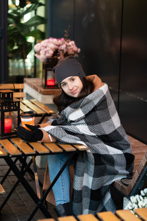 Enjoying a cozy moment on a cool autumn day with a warm drink, wrapped in a soft blanket at a charming urban cafe adorned with flowers and gentle lantern lightの写真素材