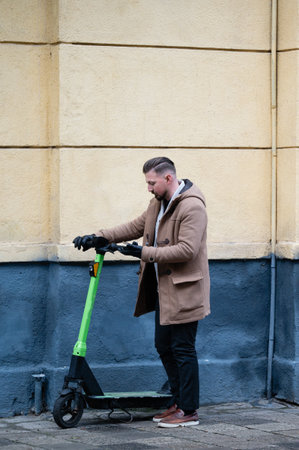 A young man dressed in a stylish brown coat stands next to a vibrant green electric scooter, inspecting it while leaning against a textured urban wall in cool weather.の写真素材