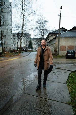 Rain dampens the pavement as a man strolls with purpose through a modest urban neighborhood, surrounded by somber buildings and muted nature in late afternoon light.の写真素材