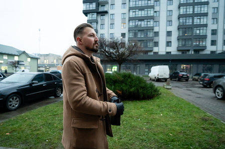 Dressed warmly, a man stands in a contemporary neighborhood during the evening, taking in the sights while surrounded by parked cars and modern buildings.の写真素材
