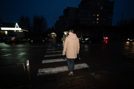 A person strides confidently across a crosswalk under the muted glow of streetlights, surrounded by vehicles moving slowly on a rainy evening in a calm urban area.の写真素材