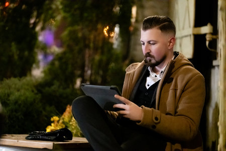 Engaged in reading or browsing, a well-dressed man comfortably sits on a ledge outside, surrounded by greenery and soft ambient light during a serene evening.の写真素材